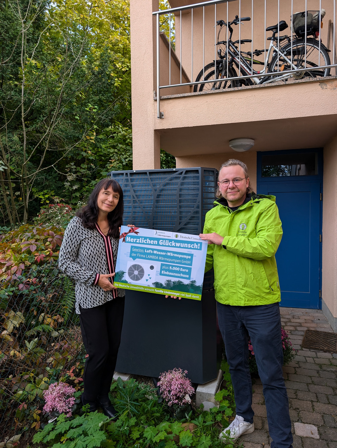 Eine Frau und ein Mann stehen vor einer neuen Wärmepumpe im Garten eines Hauses und halten ein Schild hoch, auf dem der Gewinn der Wärmepumpe draufsteht