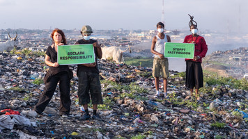 Vier Menschen stehen inmitten von Textilmüll auf einem Hügel über einem Hafen. Sie halten jeweils zu zweit Schilder hoch auf denen steht: Reclaim Responsibility und Stoppt Fast Fashion.